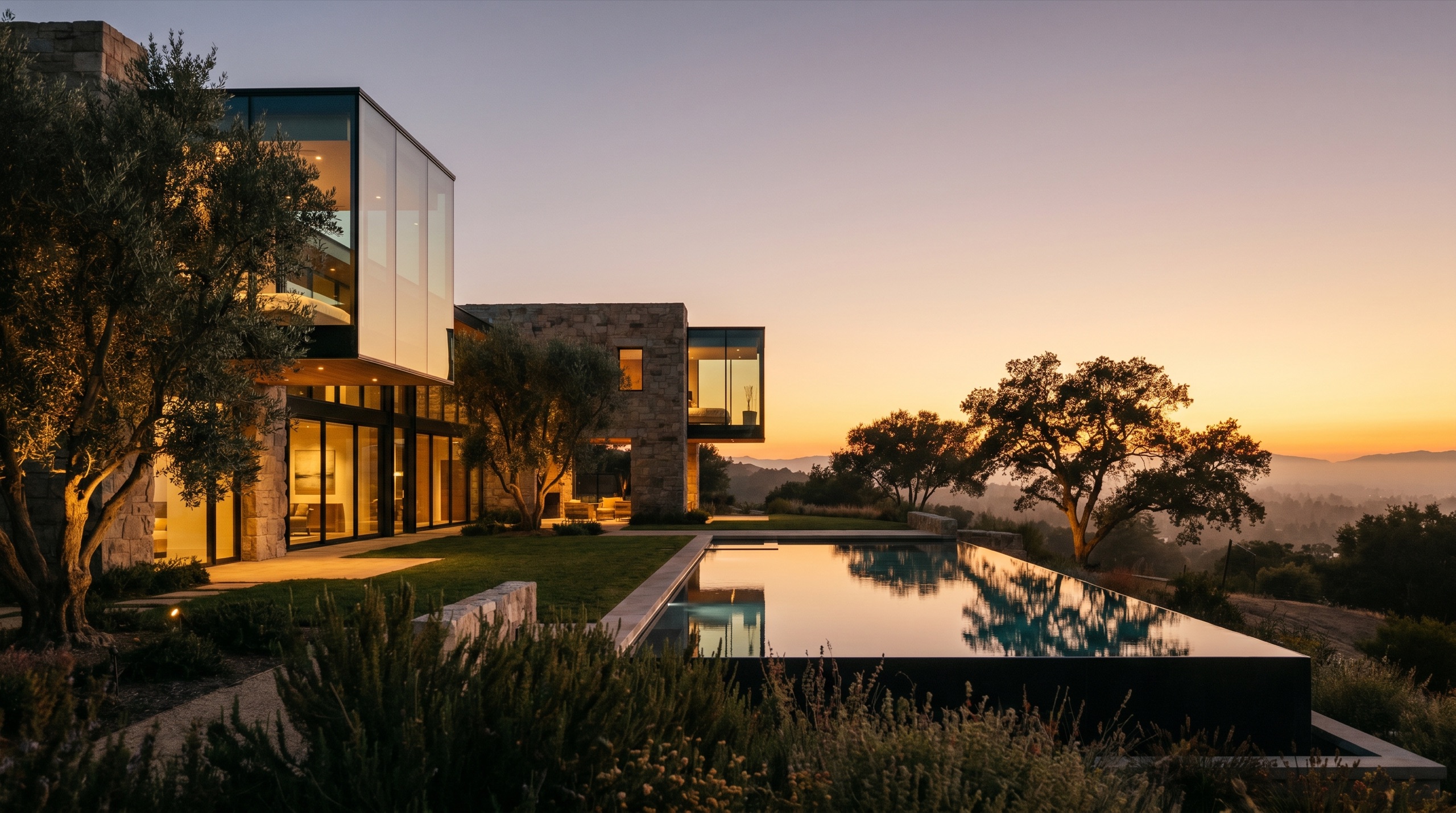 A modern luxury custom home set into the California hills at golden hour, glass and stone volumes above a still reflecting pool